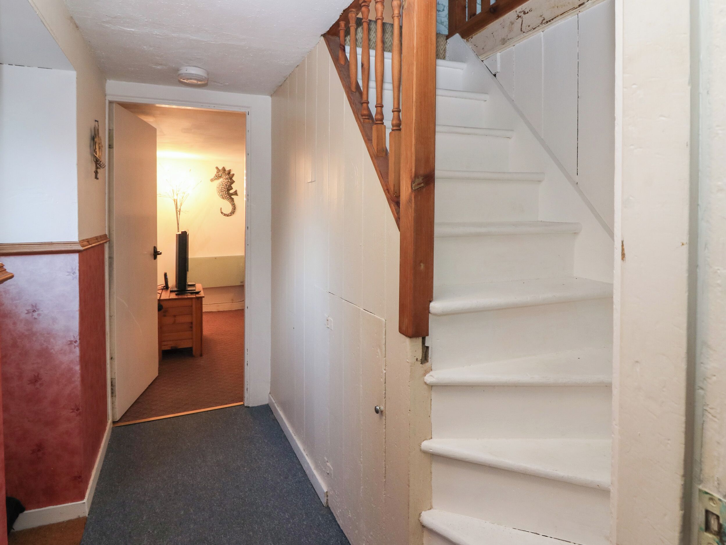 Un pasillo estrecho con una escalera de madera a la derecha, que lleva a una habitación con una puerta al final.