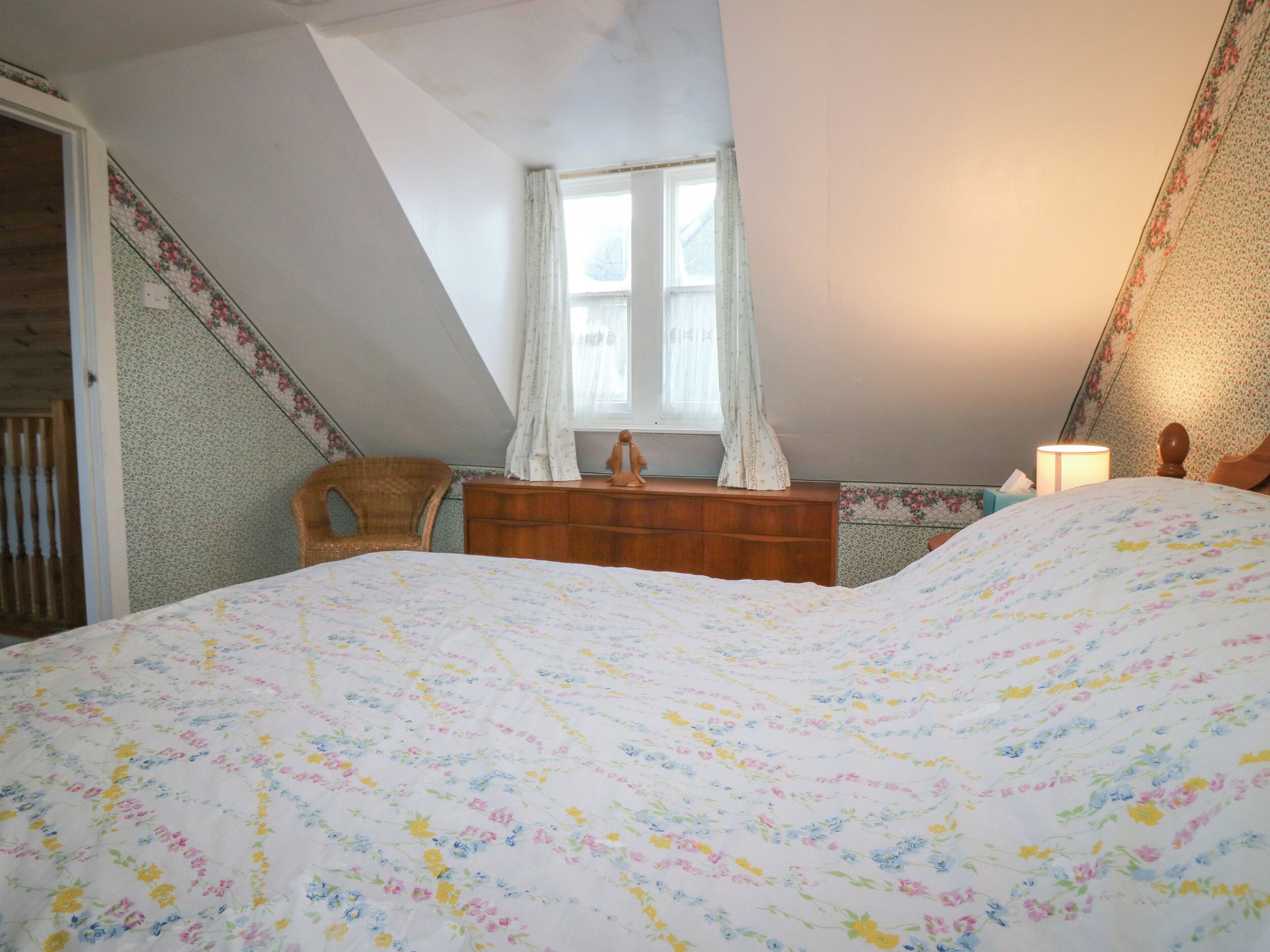 Une chambre avec un plafond en pente, un lit recouvert d’une couvre-lit à motifs floraux, une commode en bois, une chaise et une fenêtre avec des rideaux.