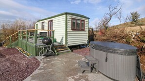 A small, single-story house with a green exterior and red-framed windows. It features a patio with a hot tub, a barbecue grill, and a small table. - Nŷth Y Bioden (Caernarfon)