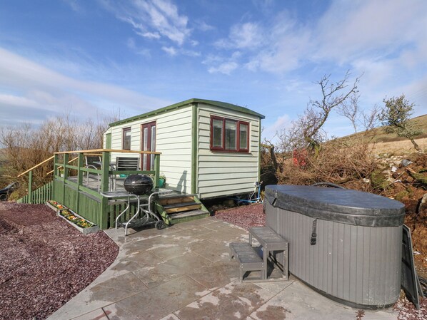Una casa pequeña de un solo piso con fachada verde y ventanas de marco rojo. Cuenta con un patio que incluye un jacuzzi, una parrilla y una pequeña mesa.