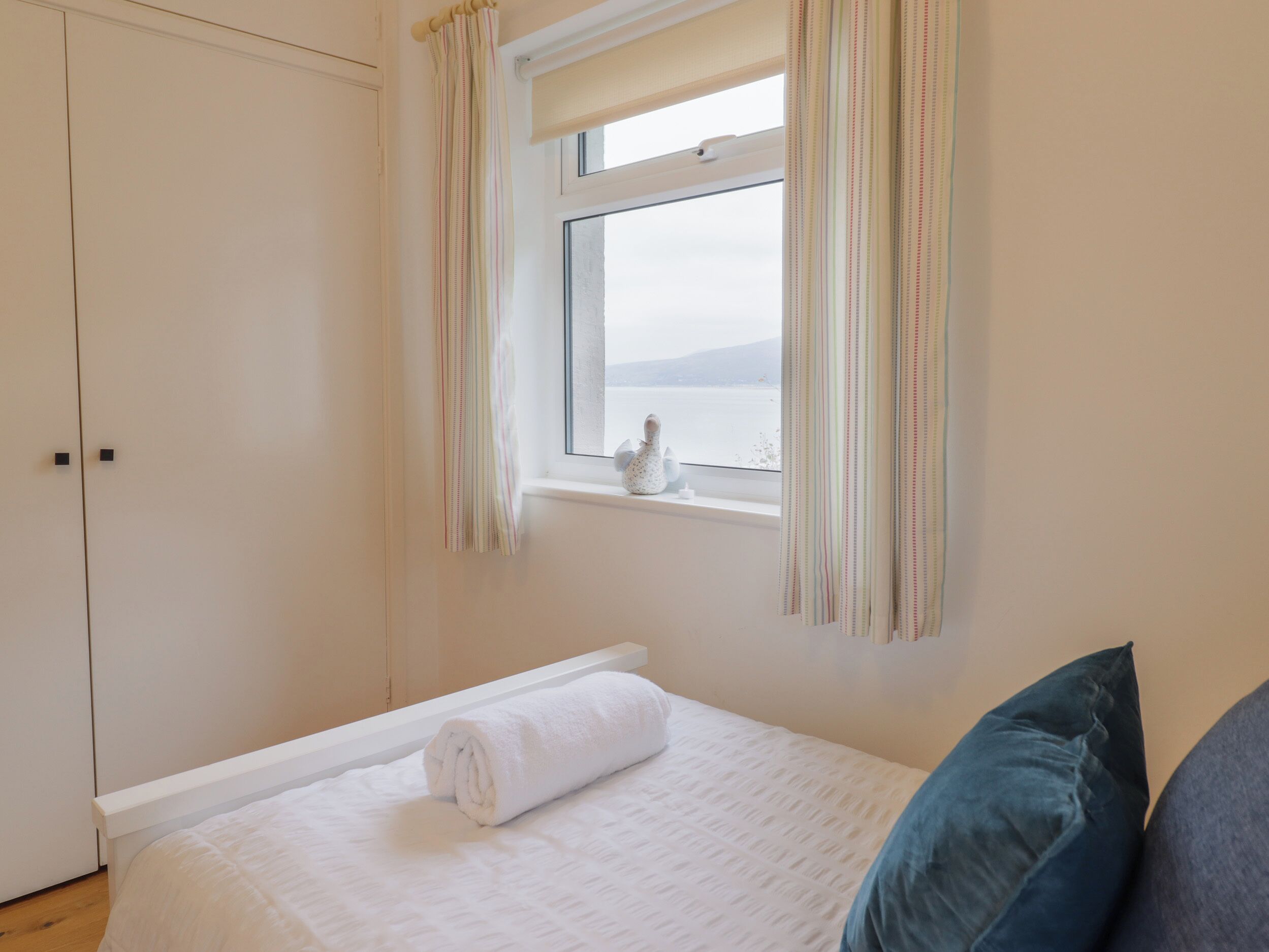A bedroom with a bed, a window with curtains, a view of the sea, and a wardrobe.