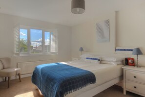 A bedroom with a large bed, a chair, a nightstand, a lamp, and a window with blinds. - 2 Studley Gardens (Lyme Regis)