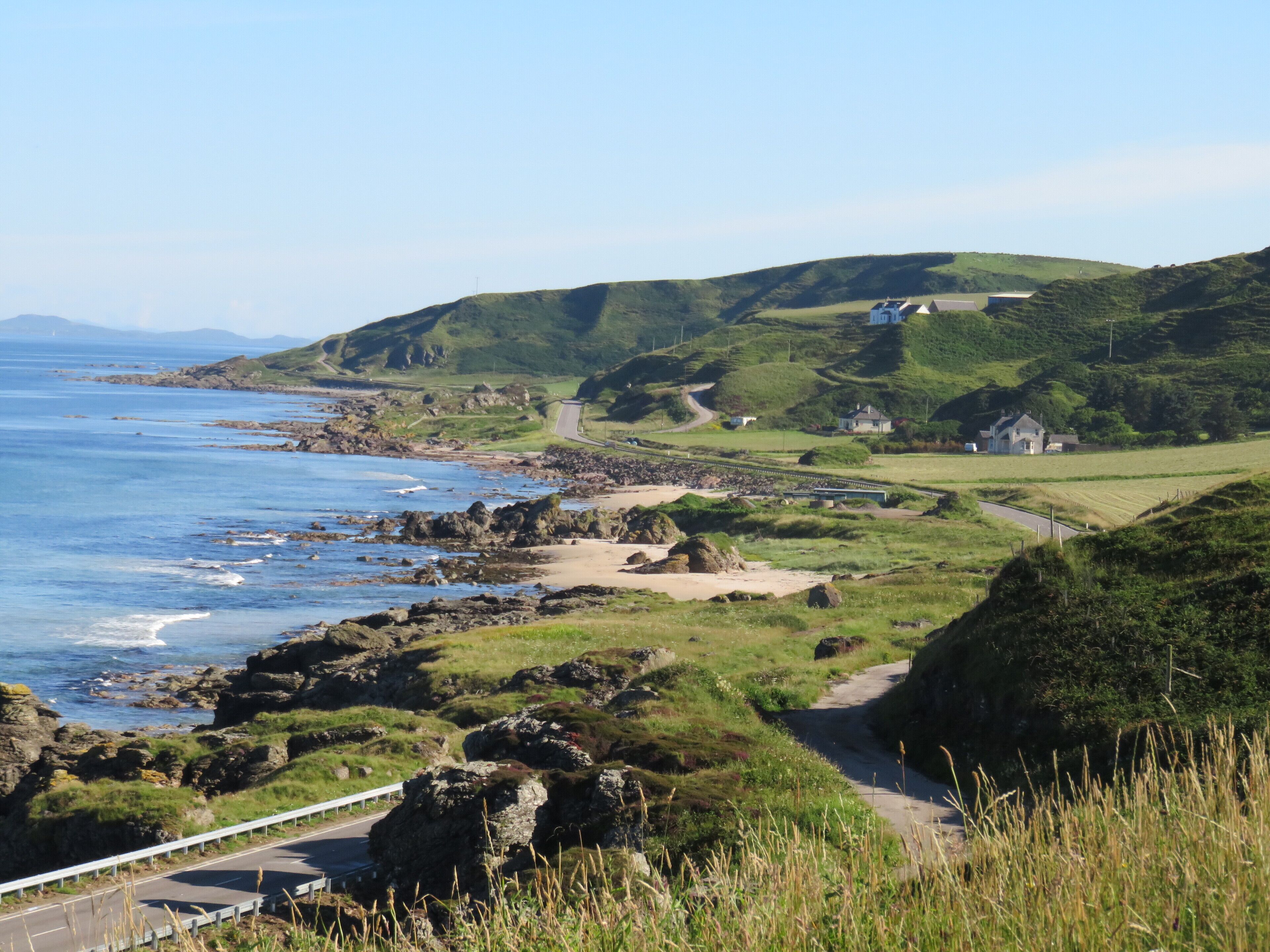 A coastal road with a guardrail, rocky shoreline, grassy hills, and a few houses in the distance.