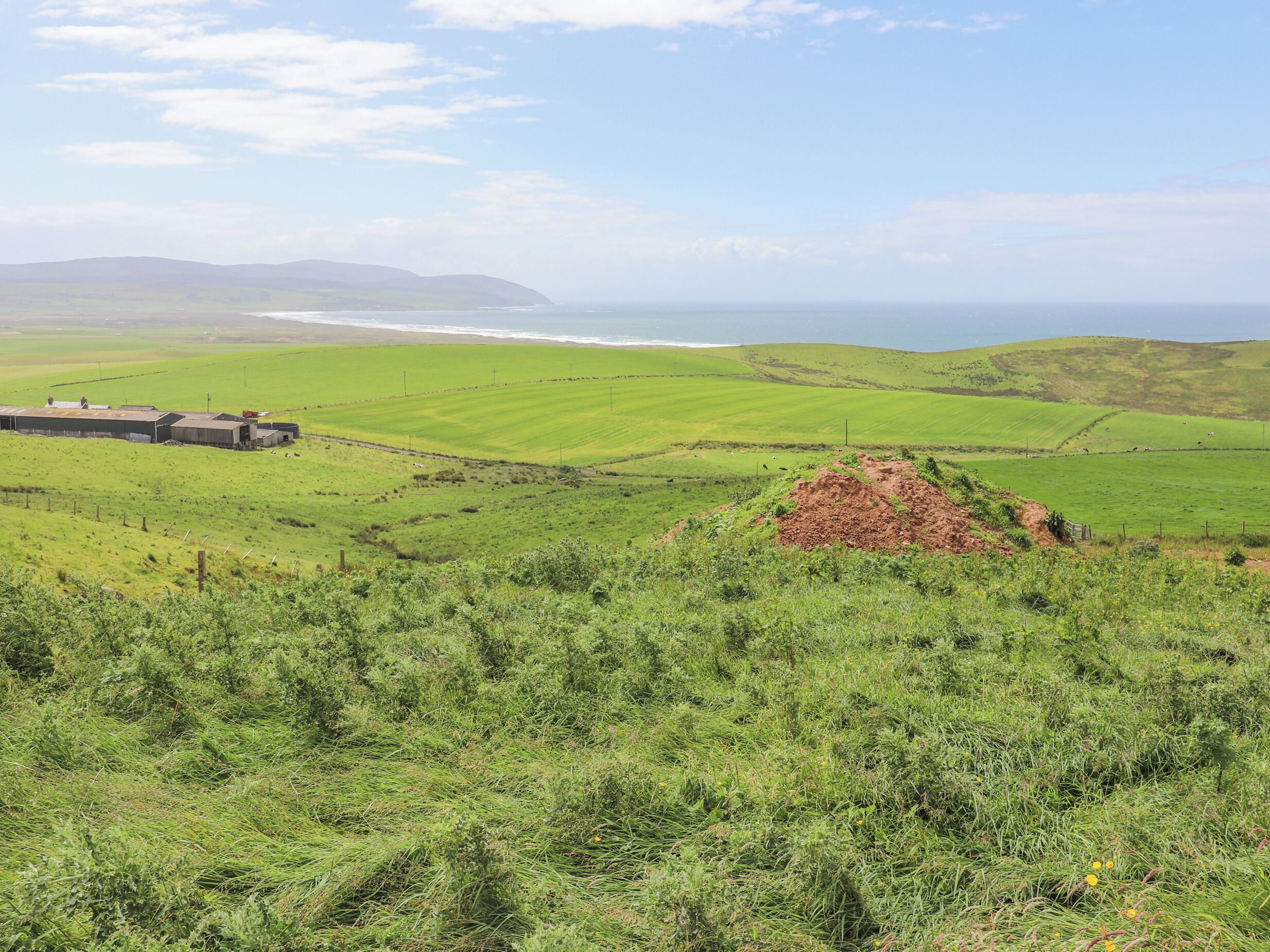 A rural landscape with green fields, a small building, and a distant view of the sea.
