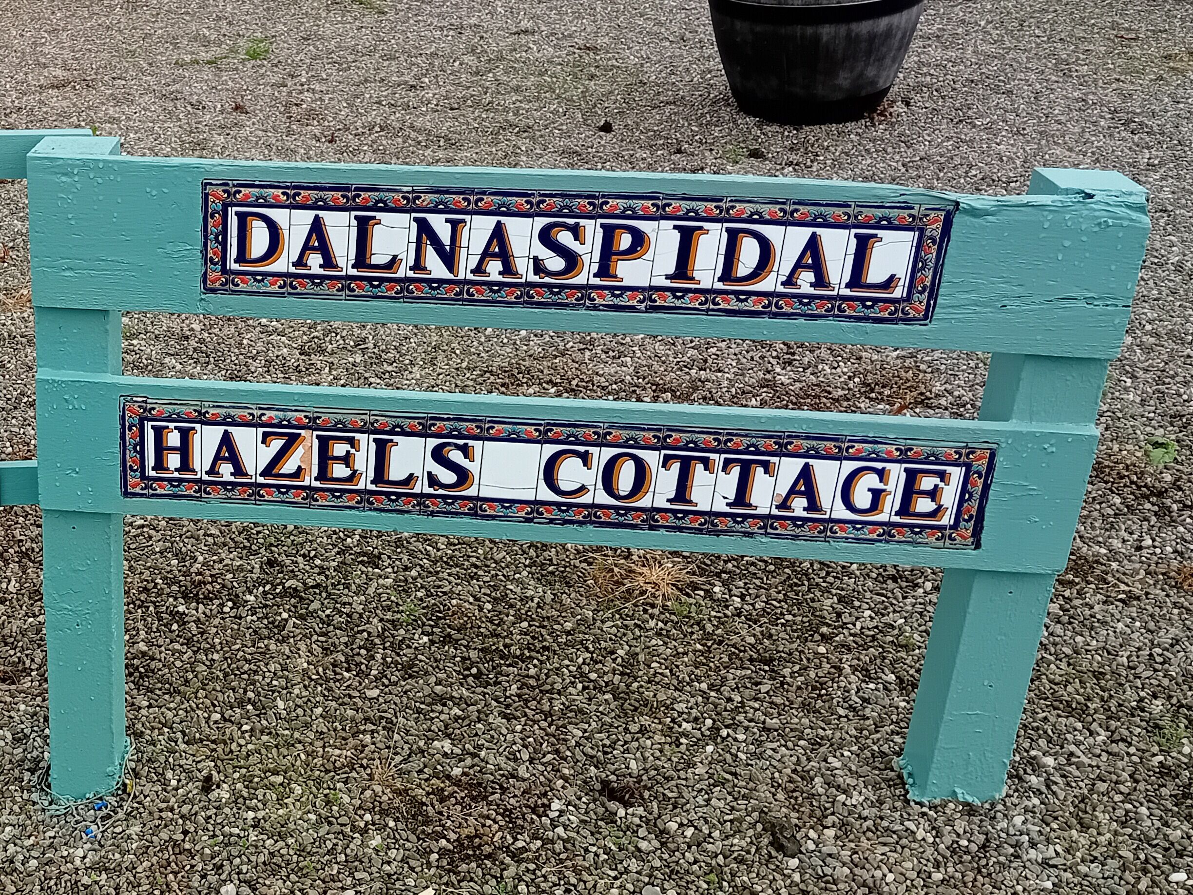 A wooden bench with two signs: "DALNASPIDAL" and "HAZELS COTTAGE".
