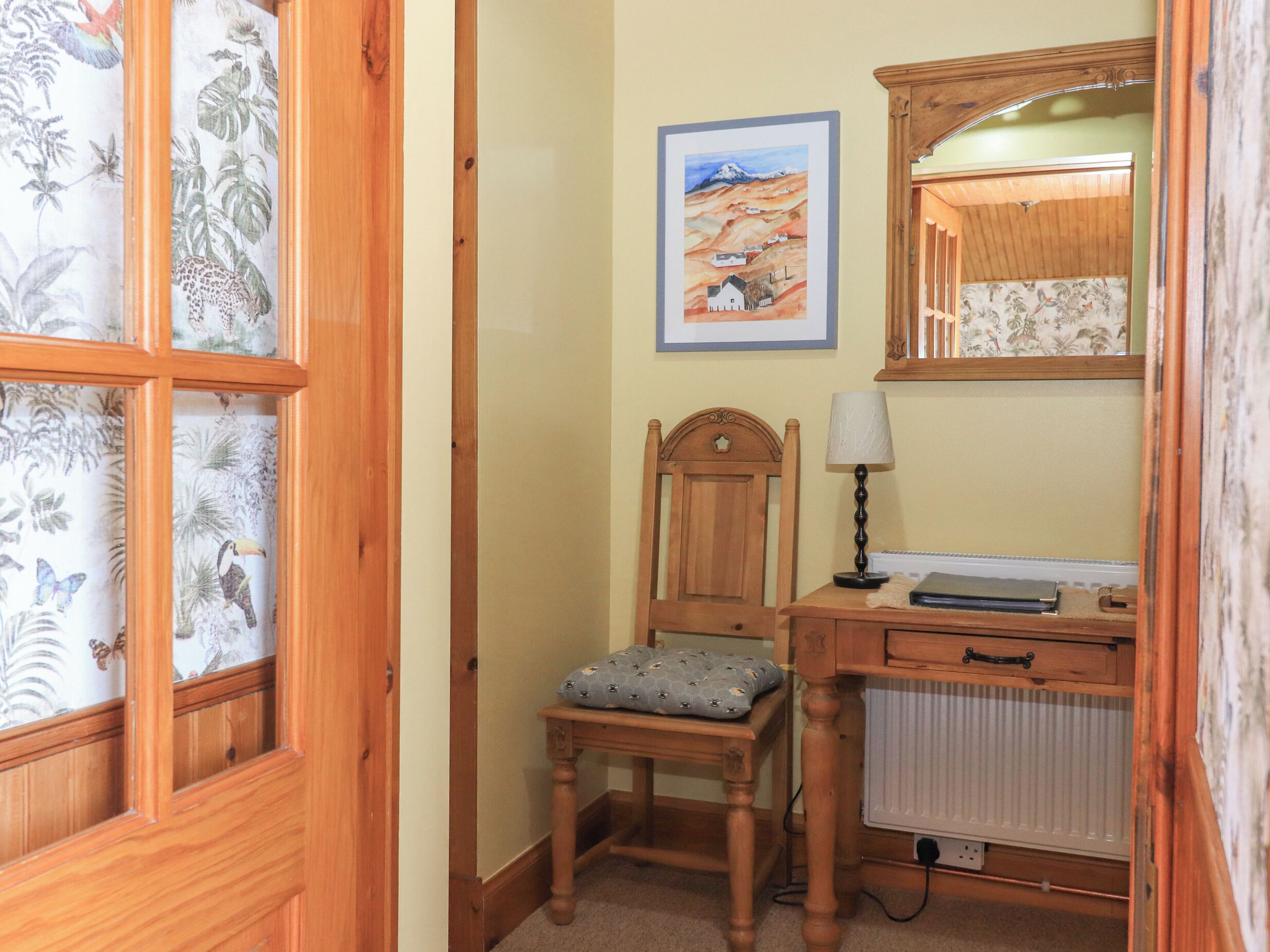 A wooden chair with a cushion, a desk with a lamp, a radiator, and a framed painting on the wall.