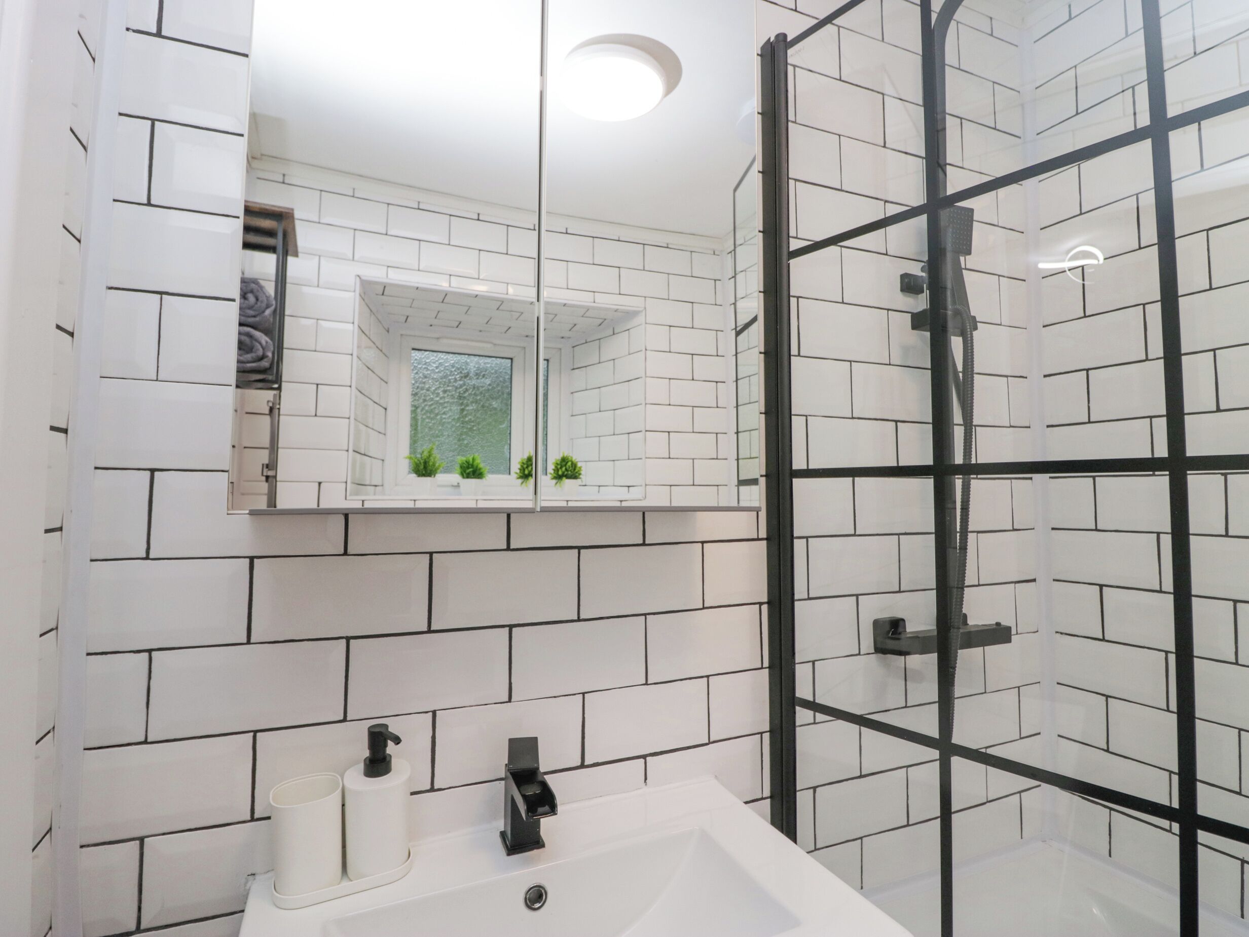 Un baño moderno con azulejos blancos tipo metro, cabina de ducha de vidrio, lavamanos blanco con dos grifos y un espejo sobre el lavamanos.