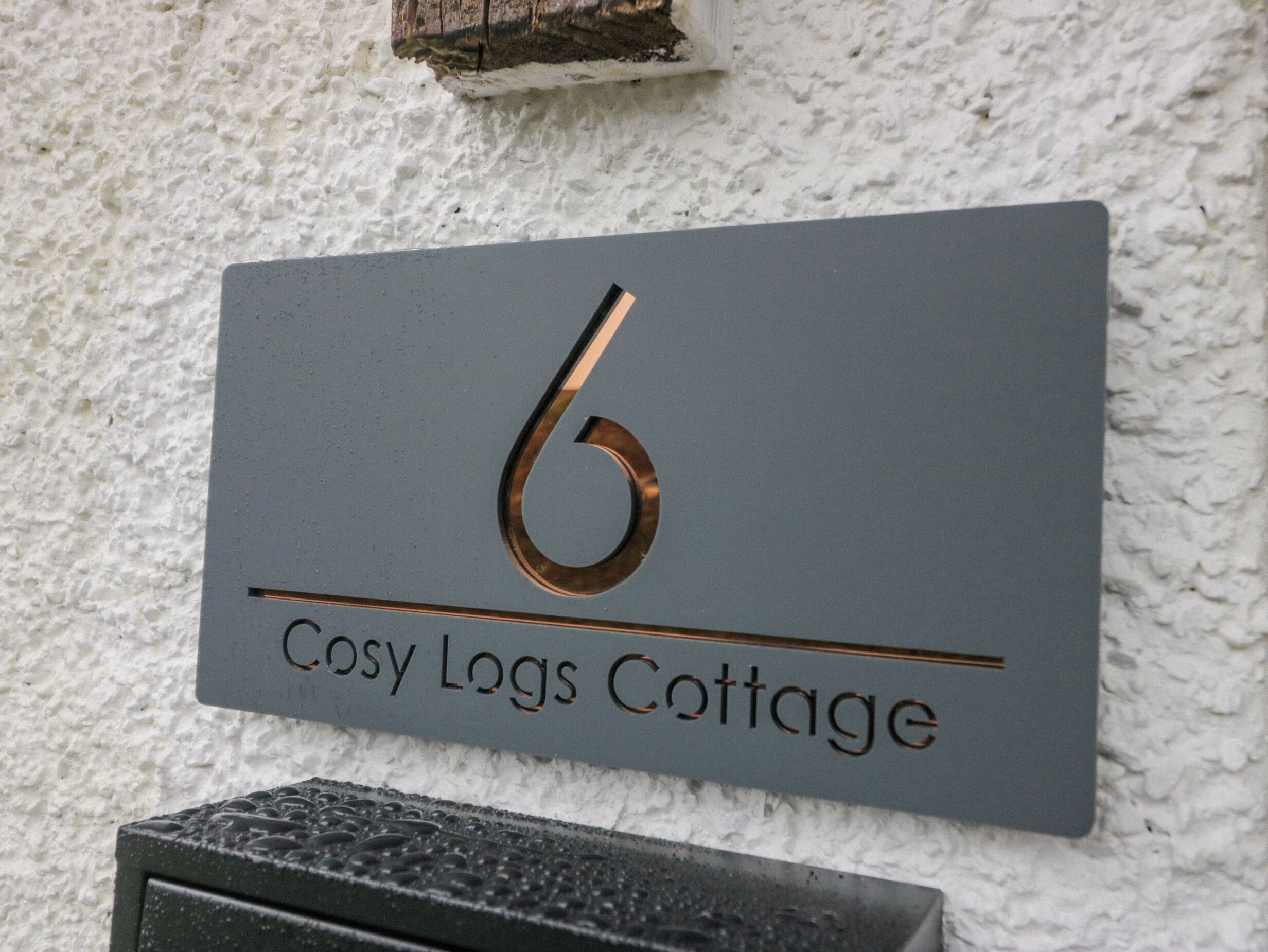 Letrero con el número de una casa, '6 Cosy Logs Cottage', fijado en una pared con textura.