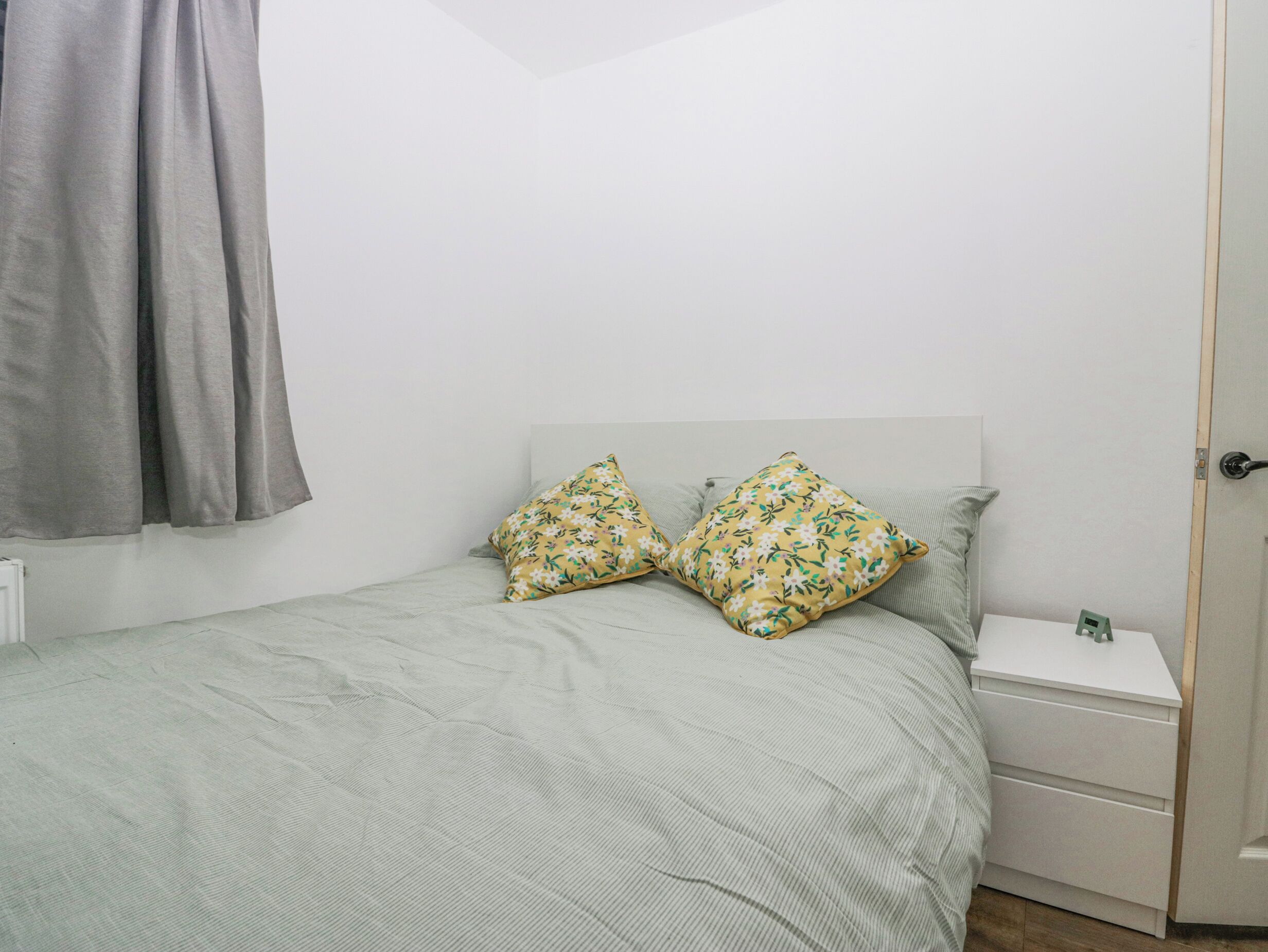 A small, neatly arranged bedroom with a bed, two pillows, a nightstand, and a door.