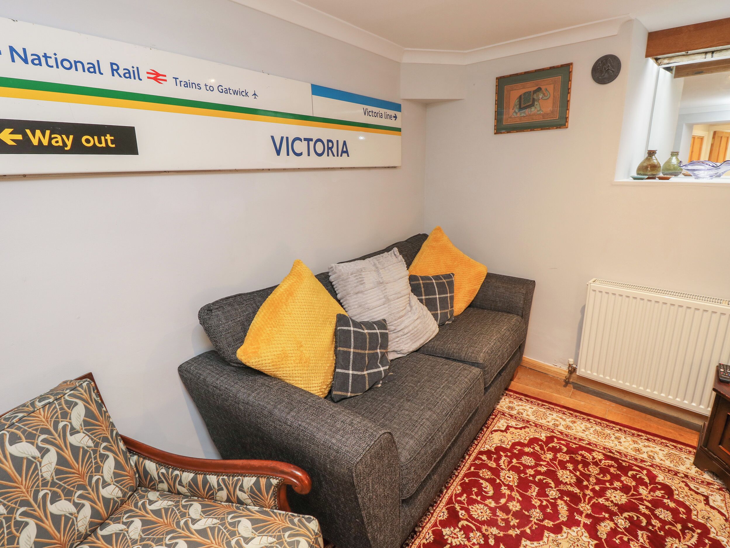 A living room with a sofa, a patterned armchair, a red and gold patterned rug, and a wall sign indicating the Victoria line.
