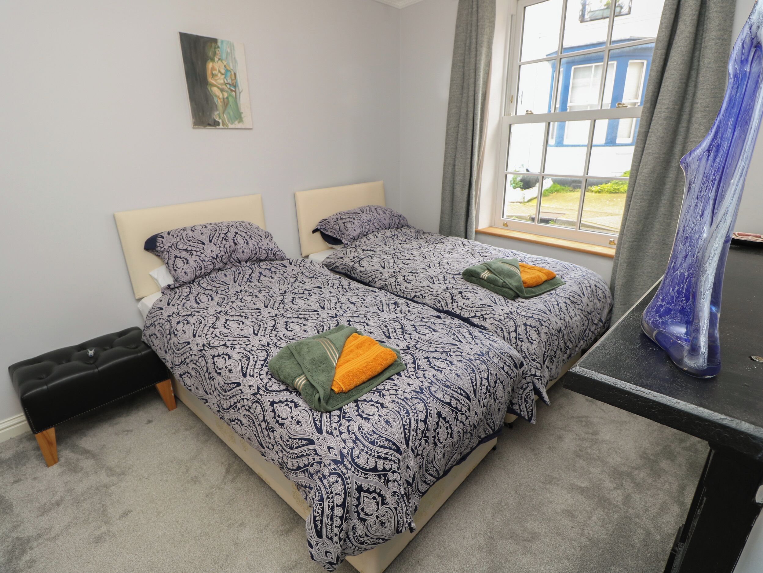 A double bed with patterned bedding, a small bedside table, a window with curtains, and a painting on the wall.