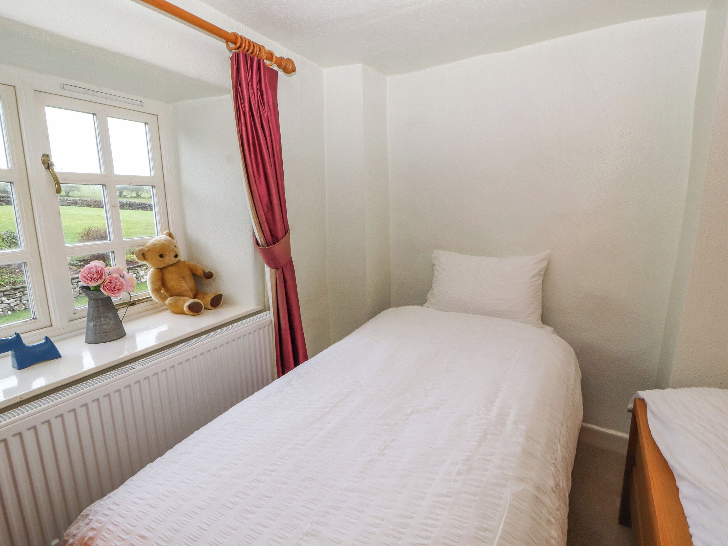 A bedroom with a single bed, a window with a view of a garden, a teddy bear, and a vase with flowers on the windowsill.