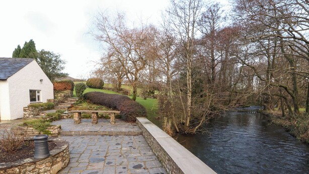 A stone pathway leading to a bench by a river, with a white building on the left. - Stang Farm (Kendal)
