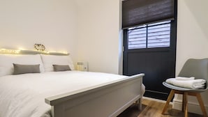 A bedroom with a bed, bedside table, chair, and a window with blinds. - Kingfisher Cottage (Canterbury)
