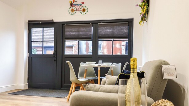 A modern living room with a sofa, a coffee table with a book and glasses, a decorative bottle, and a wall-mounted bicycle decoration. - Kingfisher Cottage (Canterbury)
