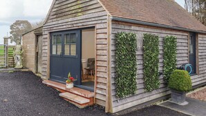 A small, single-story building with a dark door and windows, a small porch with steps, and a garden area with a statue and potted plants. - Kingfisher Cottage (Canterbury)