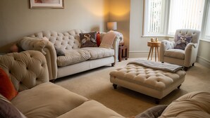 Un salon avec un canapé beige, un pouf assorti et un fauteuil assorti. On y trouve une petite table d’appoint en bois, sur laquelle sont posés une lampe et quelques objets décoratifs.
