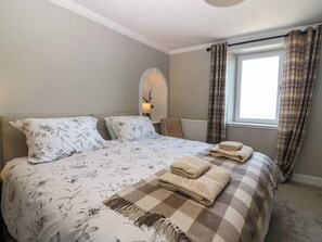A bedroom with a bed, two pillows, a bedside table with a lamp, a window with curtains, and a radiator. - West Pier Cottage (Macduff)