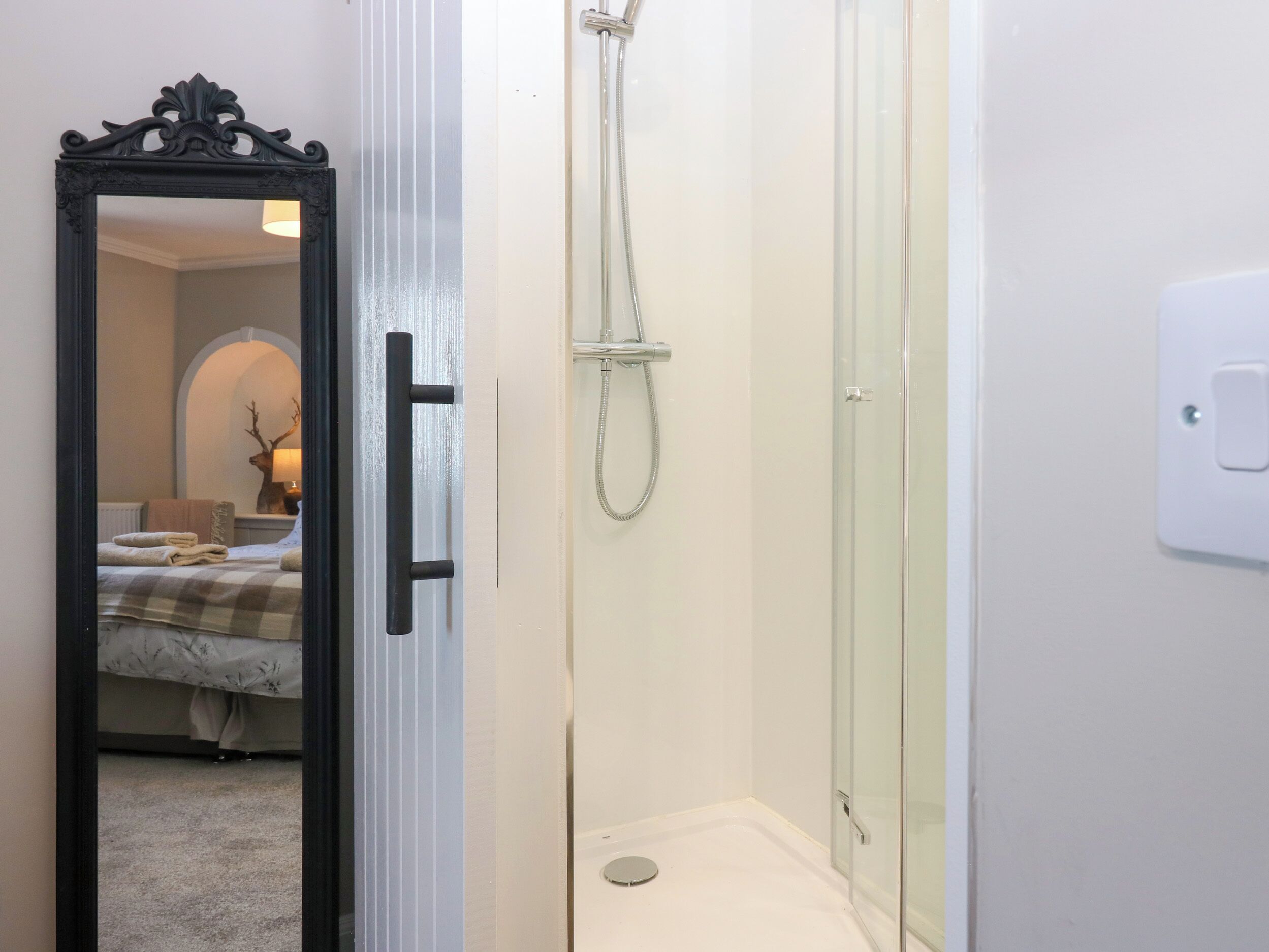 A bathroom with a glass shower enclosure, a white sink, and a mirror reflecting a bedroom with a bed and a lamp.