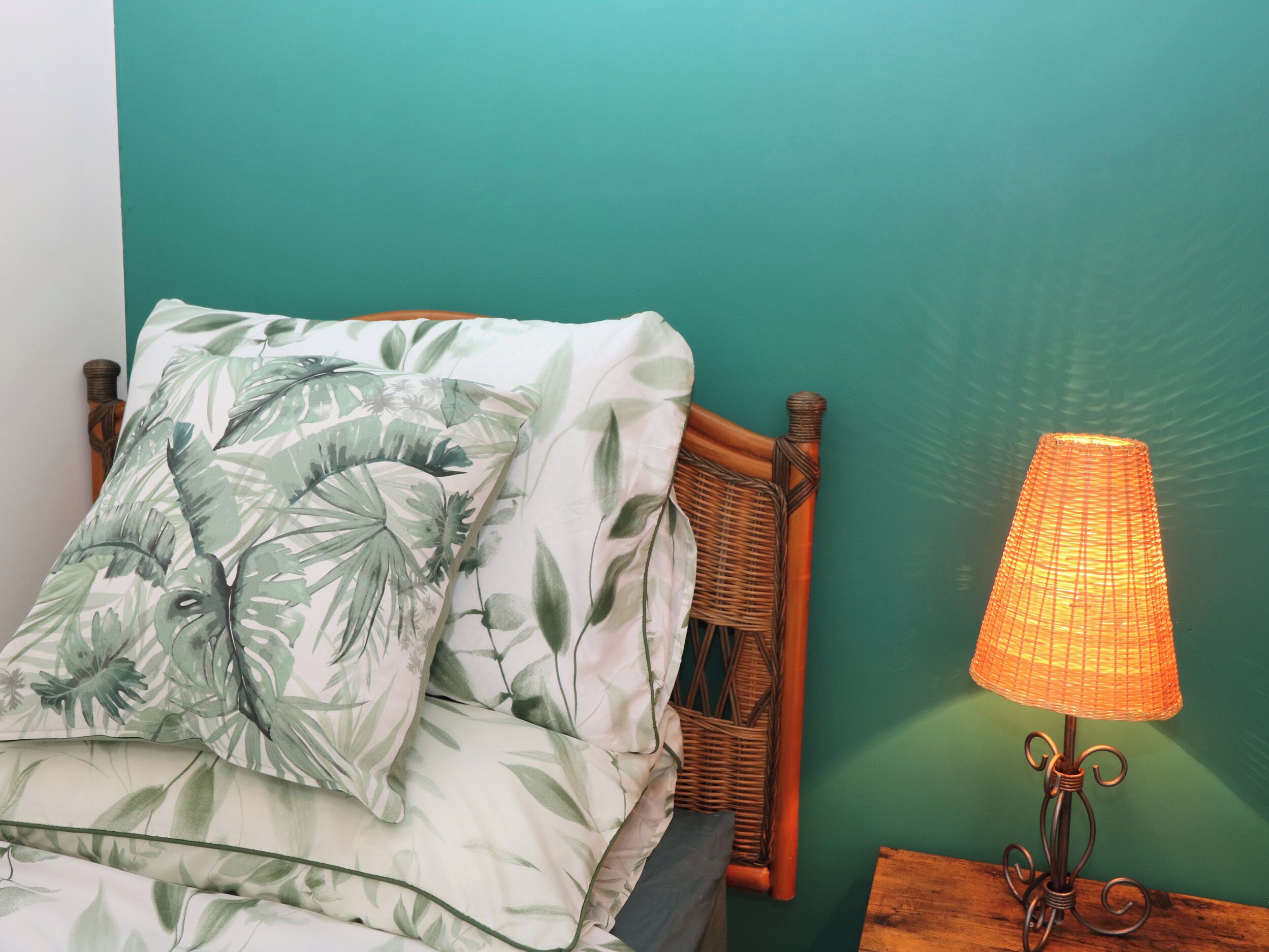 Un lit avec une couvre-lit orné de motifs tropicaux, une tête de lit en osier et une lampe de table avec un abat-jour en fibres tressées.