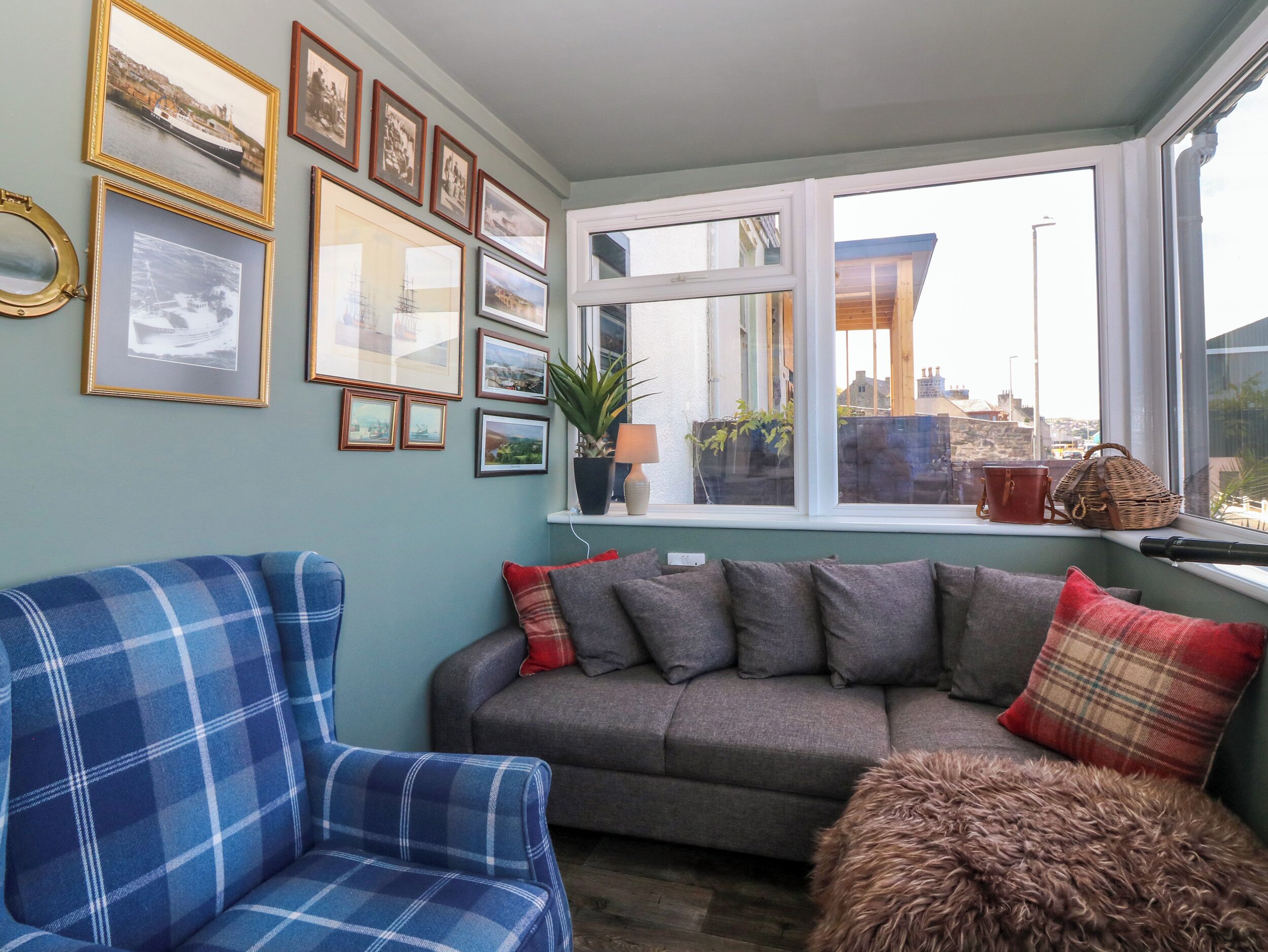 Un salón acogedor con un sillón azul de cuadros, un sofá gris con cojines rojos y de cuadros, y una ventana con vistas a edificios.
