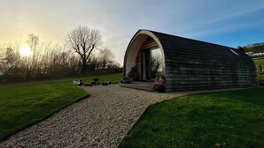 A rustic, single-story building with a curved roof, a gravel pathway leading to the entrance, and a grassy area with trees in the background. - Willow (Llanidloes)