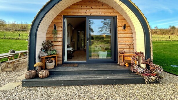 A wooden cabin with a large arched entrance, a gravel driveway, and a view of the surrounding countryside. - Willow (Llanidloes)