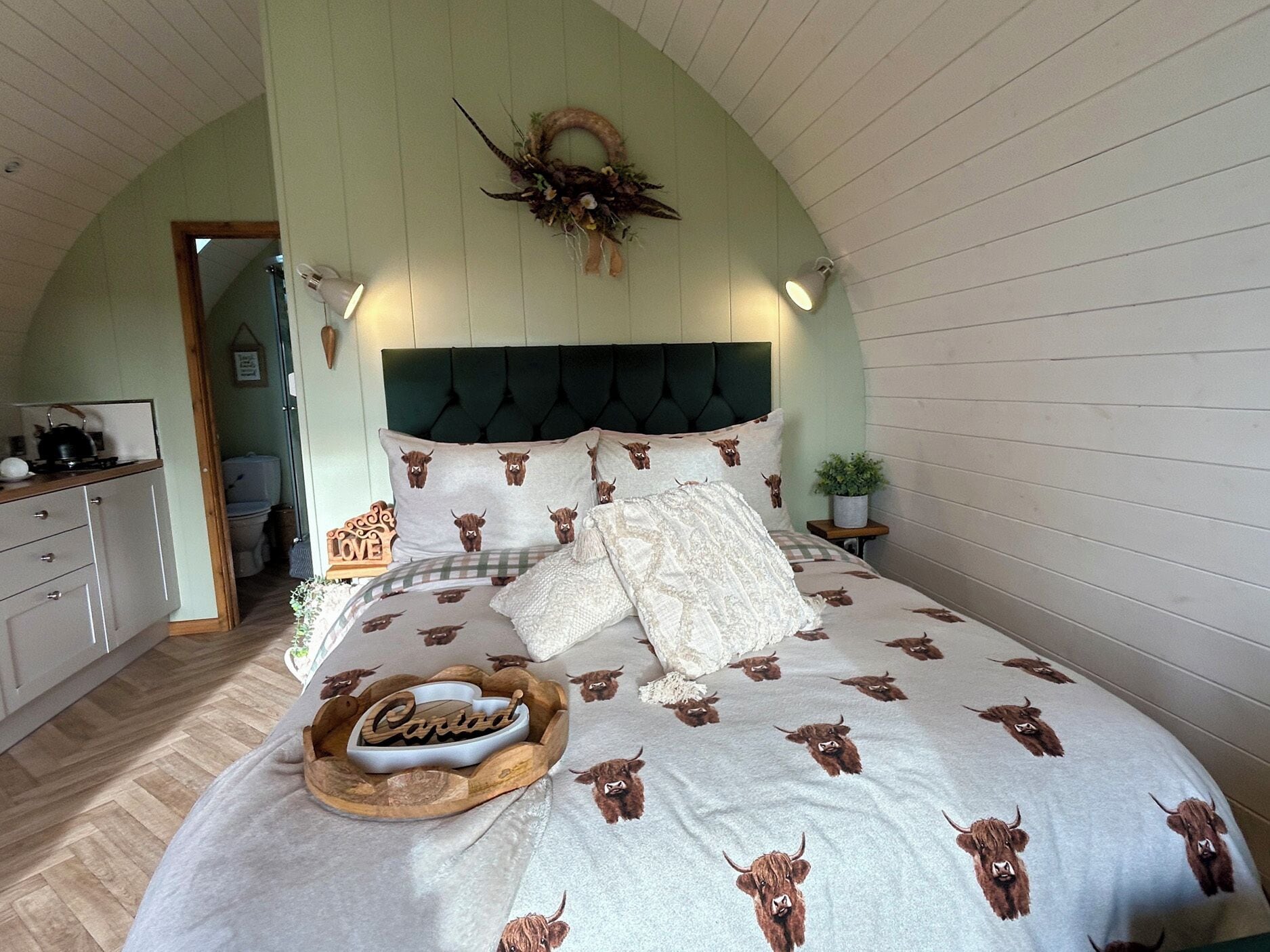 Une chambre à coucher avec un lit recouvert d’une couvre-lit à motifs de vaches, une tête de lit capitonnée, une couronne décorative accrochée au mur et une table de chevet avec une lampe et une plante.