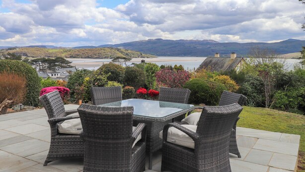 An outdoor seating area with wicker furniture and a glass table, overlooking a scenic landscape with hills and a body of water.