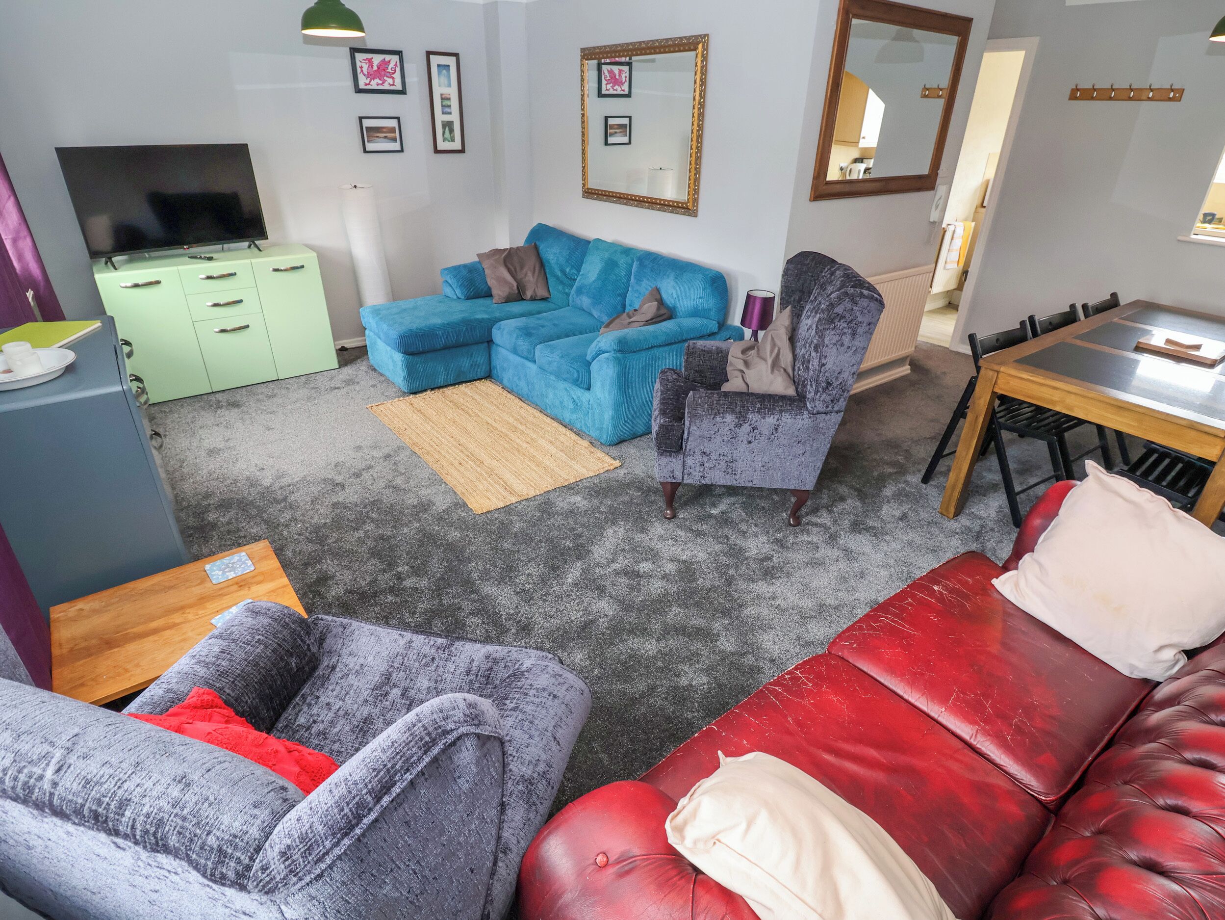 A living room with a blue sectional sofa, a grey armchair, a red leather sofa, a wooden dining table, and a green cabinet with a flat-screen TV.