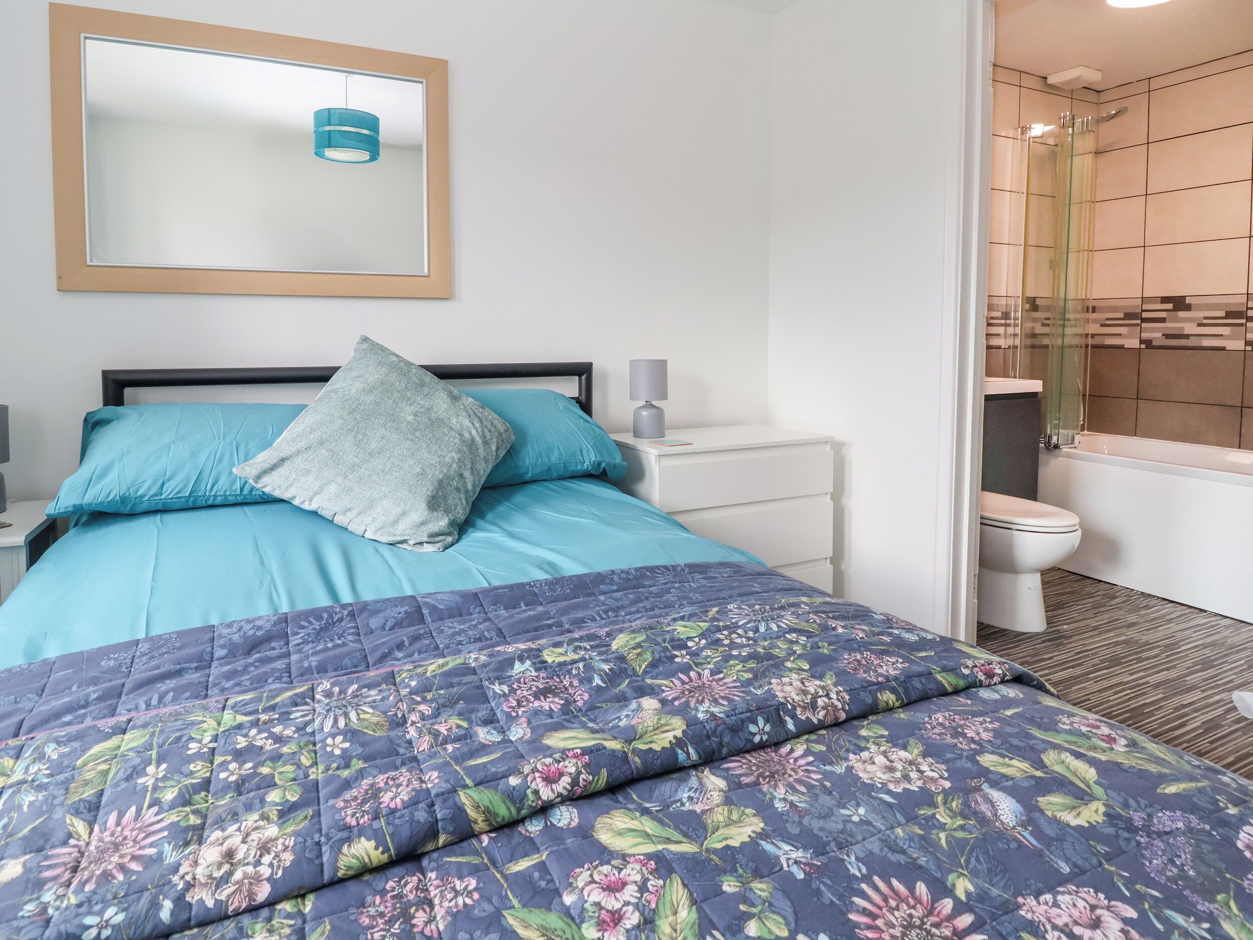 A bedroom with a bed, a bedside table, a nightstand, a mirror, and a bathroom with a toilet and shower.