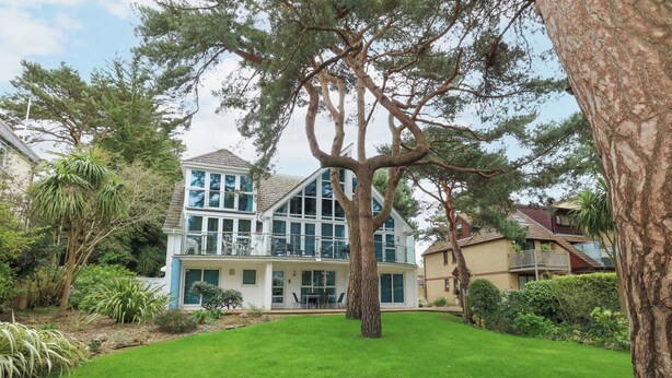 A two-story house with a balcony, surrounded by a well-maintained lawn and various trees. - 5 Red Sails (Poole)