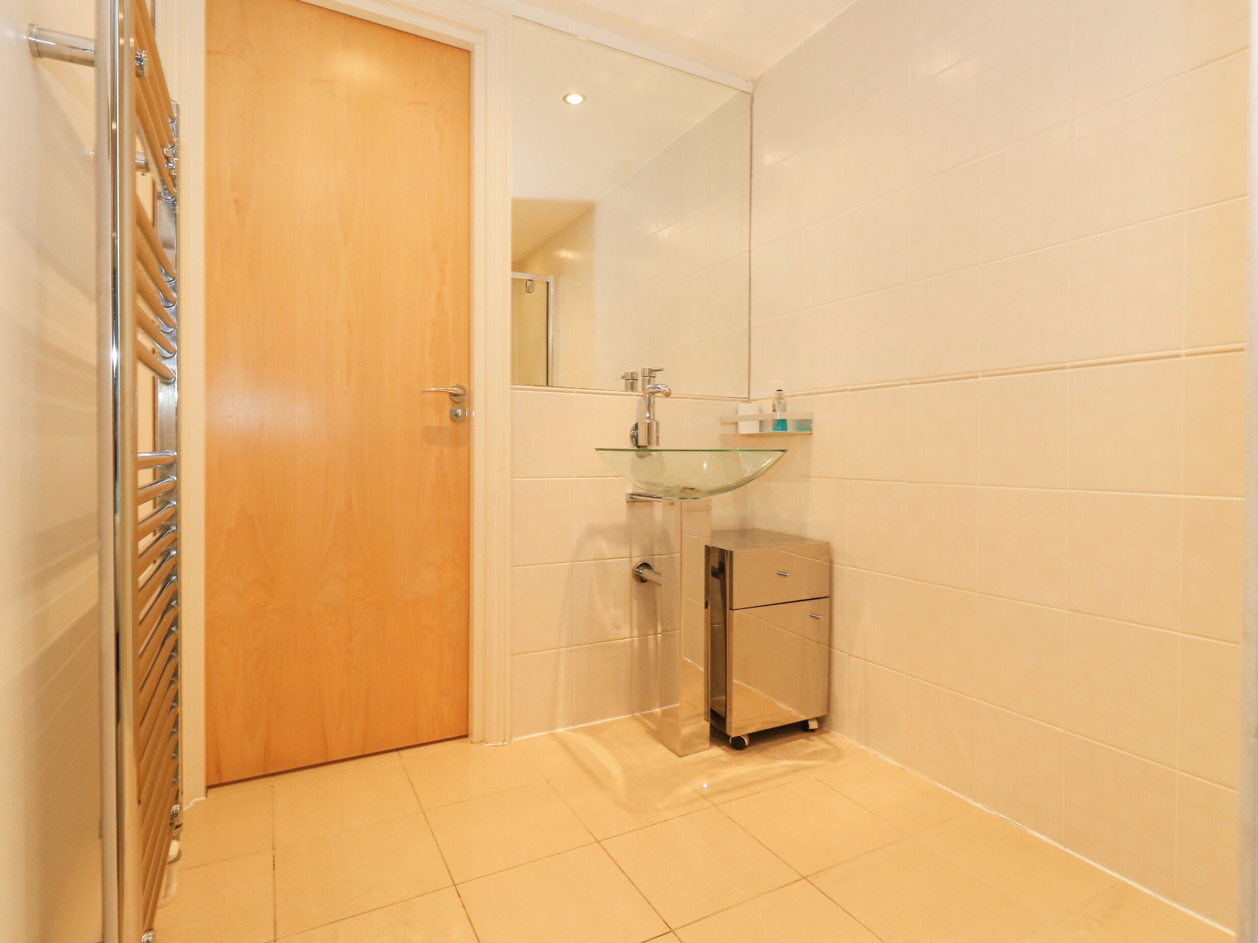Un baño con puerta de madera, lavamanos de vidrio, espejo y un mueble pequeño.