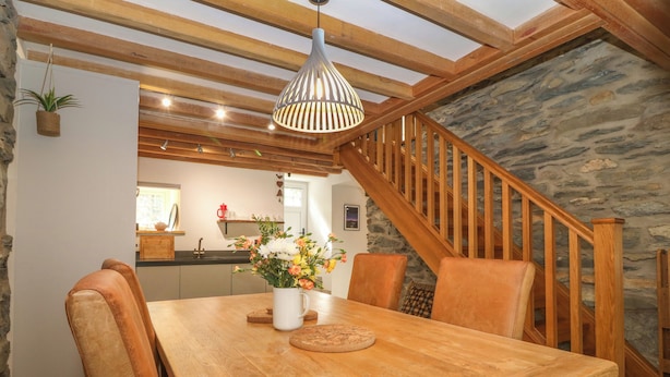 A wooden dining table with chairs, a hanging plant, and a vase of flowers. A staircase with wooden railings leads to a kitchen area. - 5 Banc Llugwy (Betws-Y-Coed)