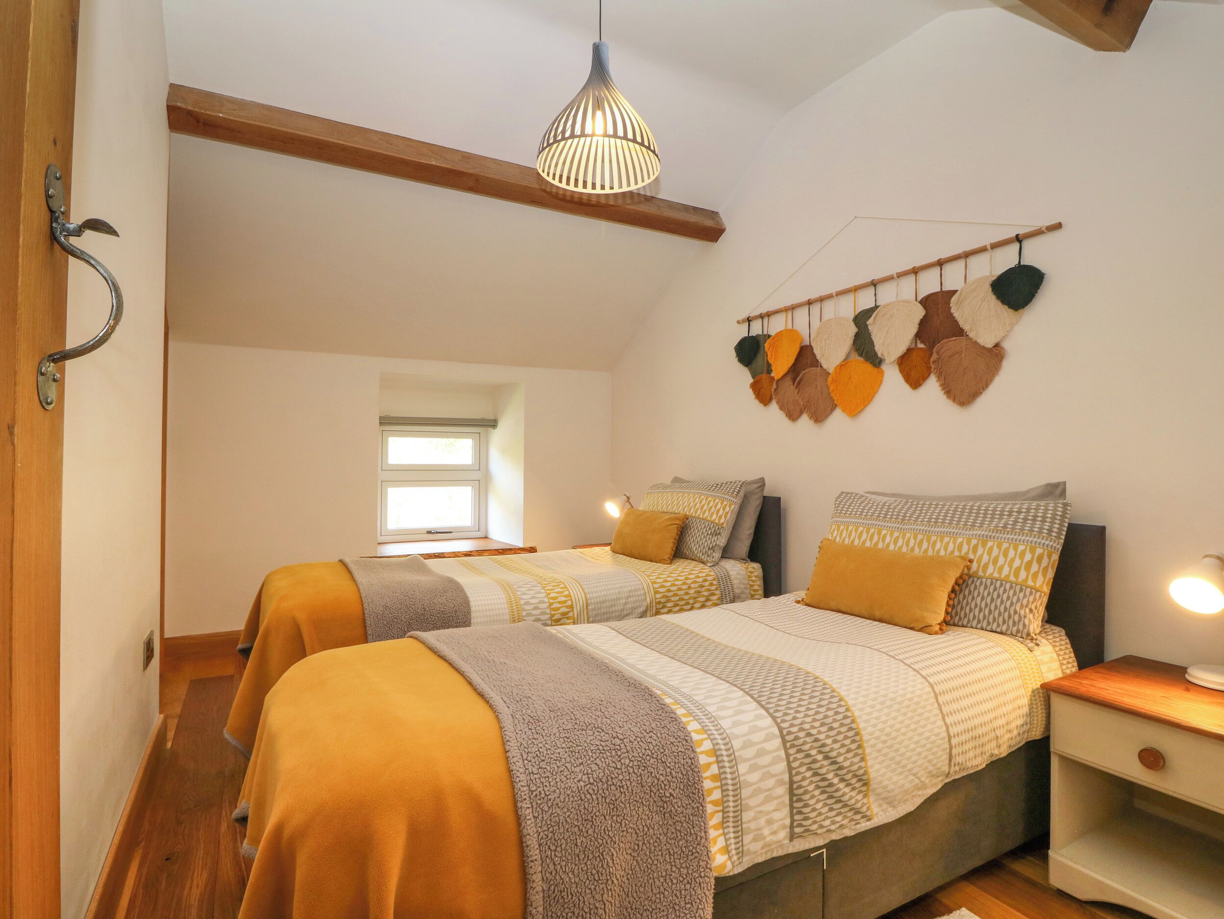 Quarto com duas camas, luminária pendente, janela e objeto decorativo na parede.