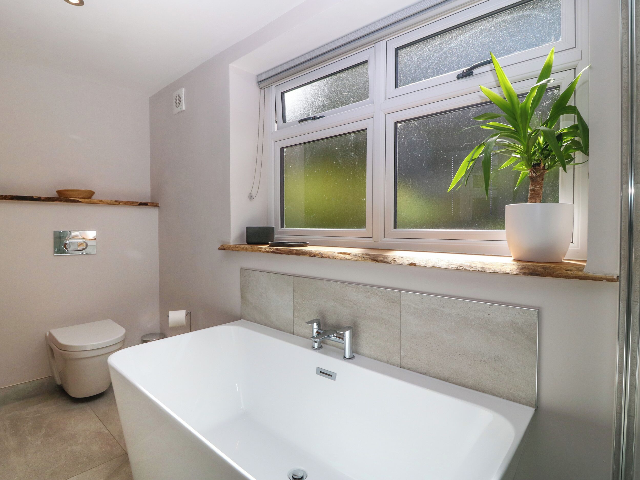Een moderne badkamer met een vrijstaand bad, een toilet en een raam met een plant op de vensterbank.