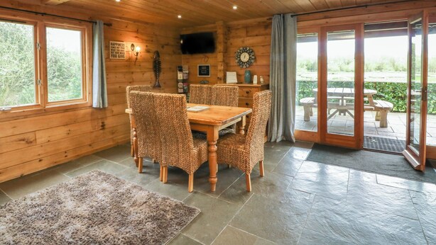 A wooden cabin interior with a dining table and chairs, a television mounted on the wall, and a view of the outdoors through a glass door. - Aspen (Winkleigh)