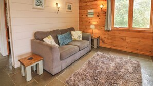 A cozy room with a wooden ceiling, a beige sofa with floral cushions, a small wooden table, a fluffy rug, and a window with curtains. - Aspen (Winkleigh)