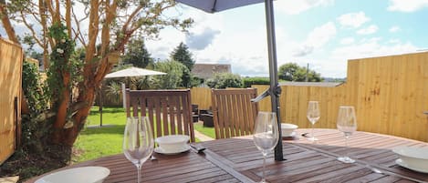 Un área de comedor al aire libre con una mesa de madera dispuesta para cuatro personas, incluyendo platos, tazones, copas de vino y cubiertos.