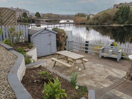 Un patio con una mesa y bancos de madera, un cobertizo y vista a un río y edificios al fondo.