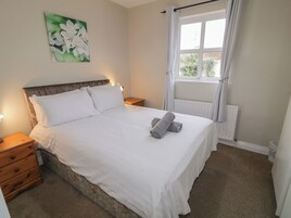Una cama bien hecha con sábanas blancas y dos toallas enrolladas, una mesita de noche de madera con una lámpara, un cuadro de flores blancas en la pared y una ventana con cortinas blancas.