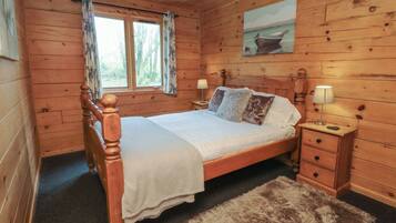 Un dormitorio de cabaña de madera con una cama, dos mesitas de noche, una lámpara de noche, una ventana con cortinas y un cuadro en la pared.