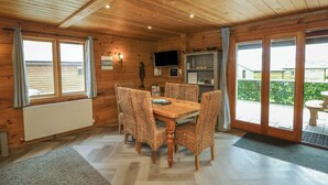 A wooden cabin with a dining area featuring a wooden table and woven chairs, a television mounted on the wall, and a view of the outdoors through large sliding glass doors. - Maple (Winkleigh)