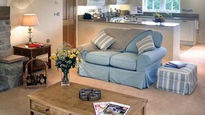 A living room with a sofa, a coffee table, and a lamp. There is a kitchen in the background with cabinets and a window. - The Lake Cottage (Belford)