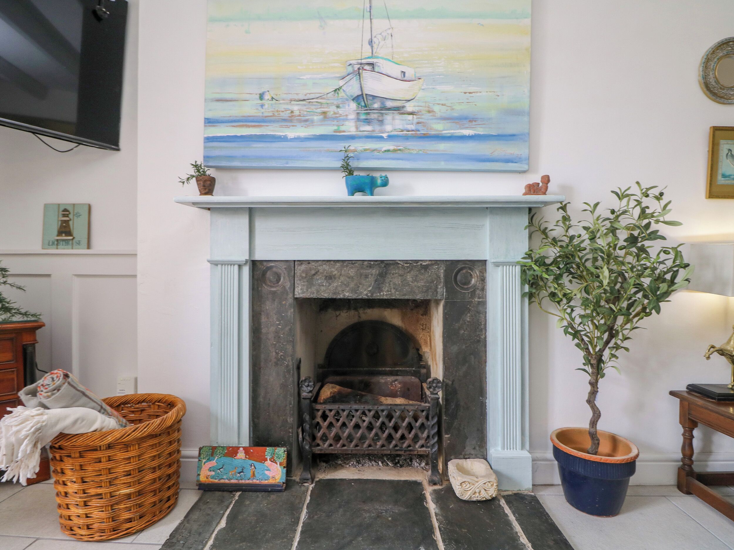 A living room with a fireplace, a painting of a sailboat, a wicker basket, and a potted plant.