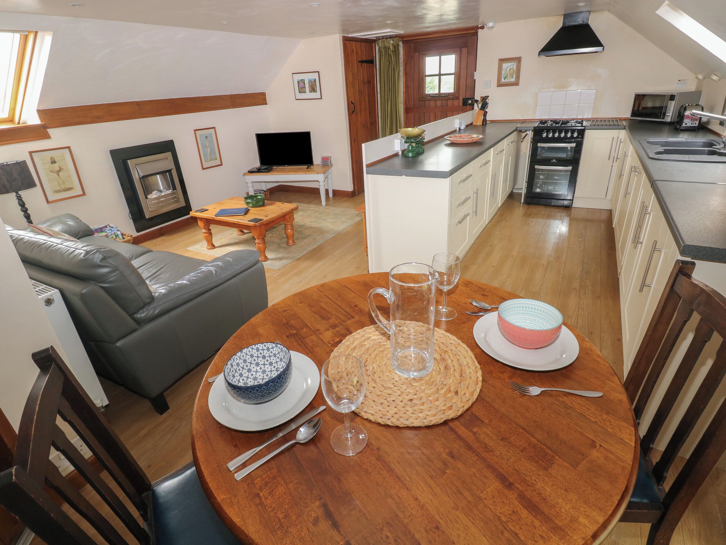 Sala de estar com sofá, mesa de jantar posta com bolo, copos e jarra. Cozinha ao fundo com micro-ondas e fogão.
