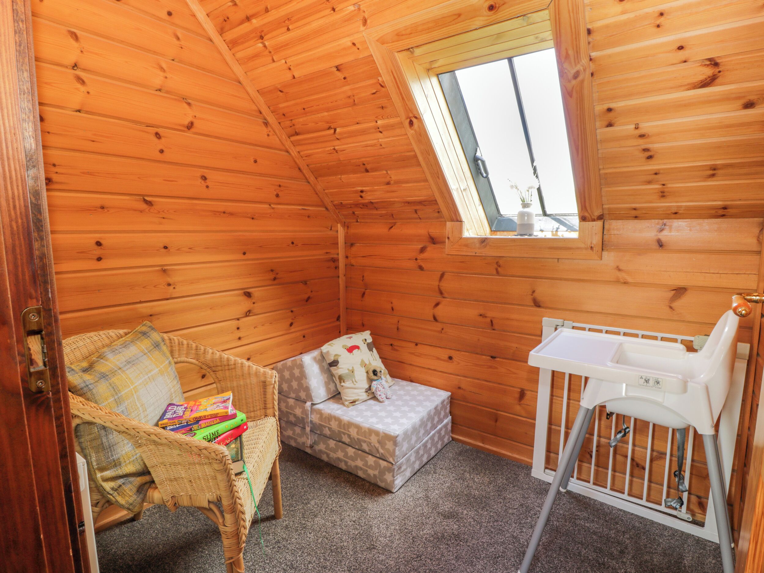 Une chambre mansardée chaleureuse avec des murs en bois, une petite fenêtre, un berceau, une chaise en osier et une table de chevet.