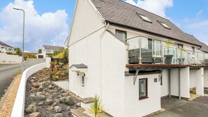 A white two-story house with a balcony, a small garden with rocks and plants, and a clear sky. - 220 Cae Du (Pwllheli)