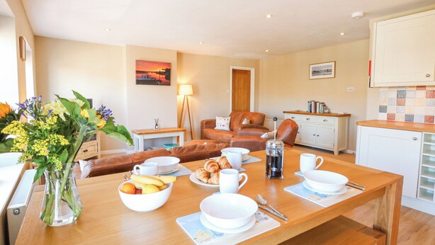 A dining area with a wooden table set for breakfast, including croissants, coffee, and fruit. There is a sofa and a chair in the background, and a kitchen area with white cabinets and a tiled backsplash. - Crab Corner (Wells-next-the-Sea)