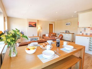 A dining area with a wooden table set for breakfast, including croissants, coffee, and fruit. There is a sofa and a chair in the background, and a kitchen area with white cabinets and a tiled backsplash. - Crab Corner (Wells-next-the-Sea)